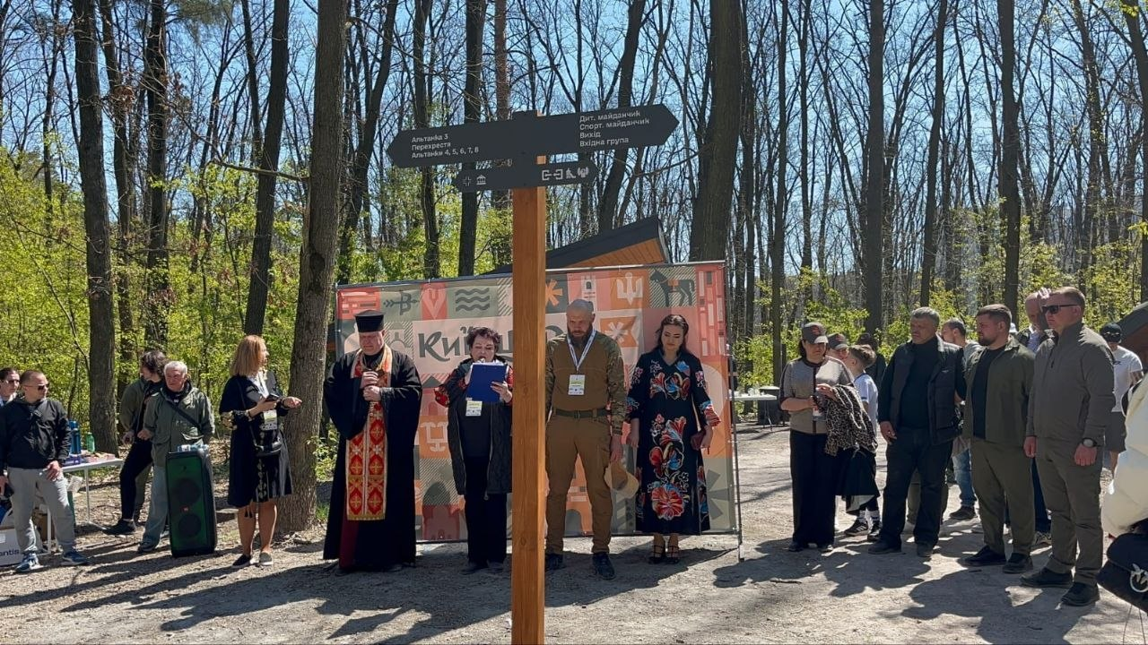 Урочистий захід у лісопарковій зоні: група людей, серед яких священник та ведучі, стоїть біля банера «Київщина» поруч із дерев'яним вказівником напрямків.