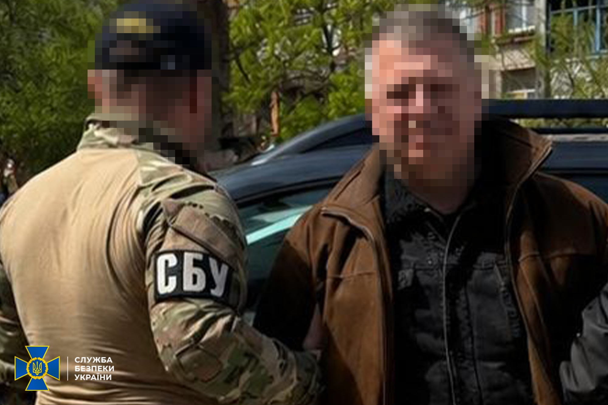Співробітник СБУ у камуфляжній формі затримує чоловіка в коричневій куртці на вулиці. Обличчя обох осіб приховані, на рукаві військового видно напис «СБУ».