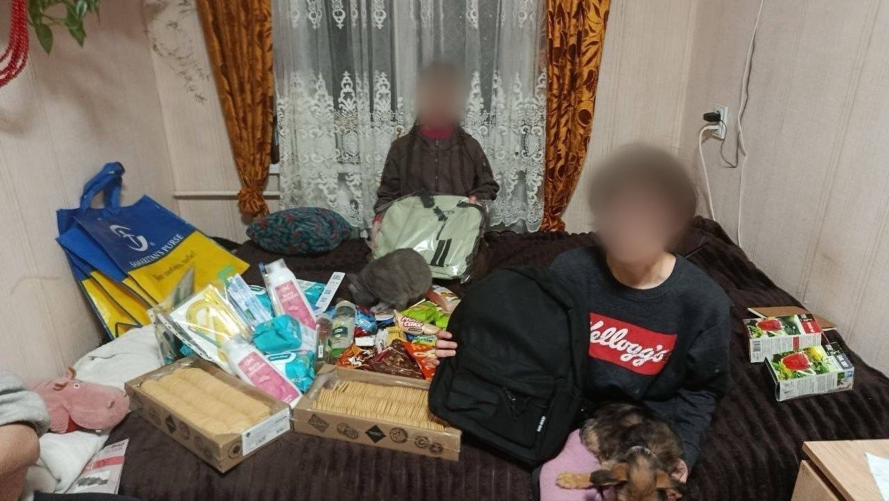 Двоє дітей із розмитими обличчями сидять на ліжку в кімнаті, оточені пакунками з гуманітарною допомогою. Перед ними лежать засоби гігієни, продукти харчування, солодощі та нові рюкзаки.