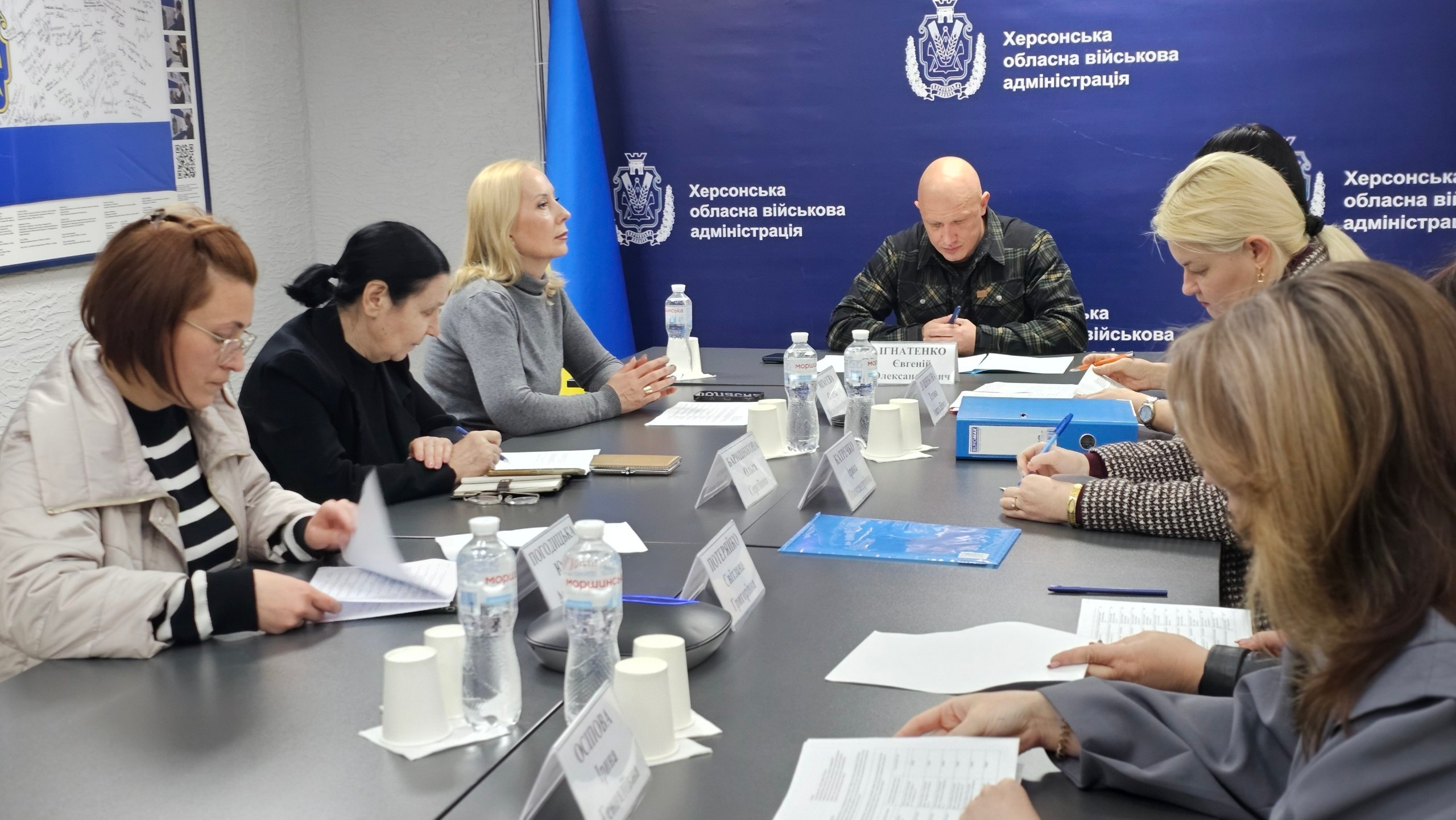 Група людей сидить за великим столом під час офіційної наради. Учасники обговорюють документи, роблять записи та слухають доповідача на фоні синього банера з державною символікою та офіційними написами.