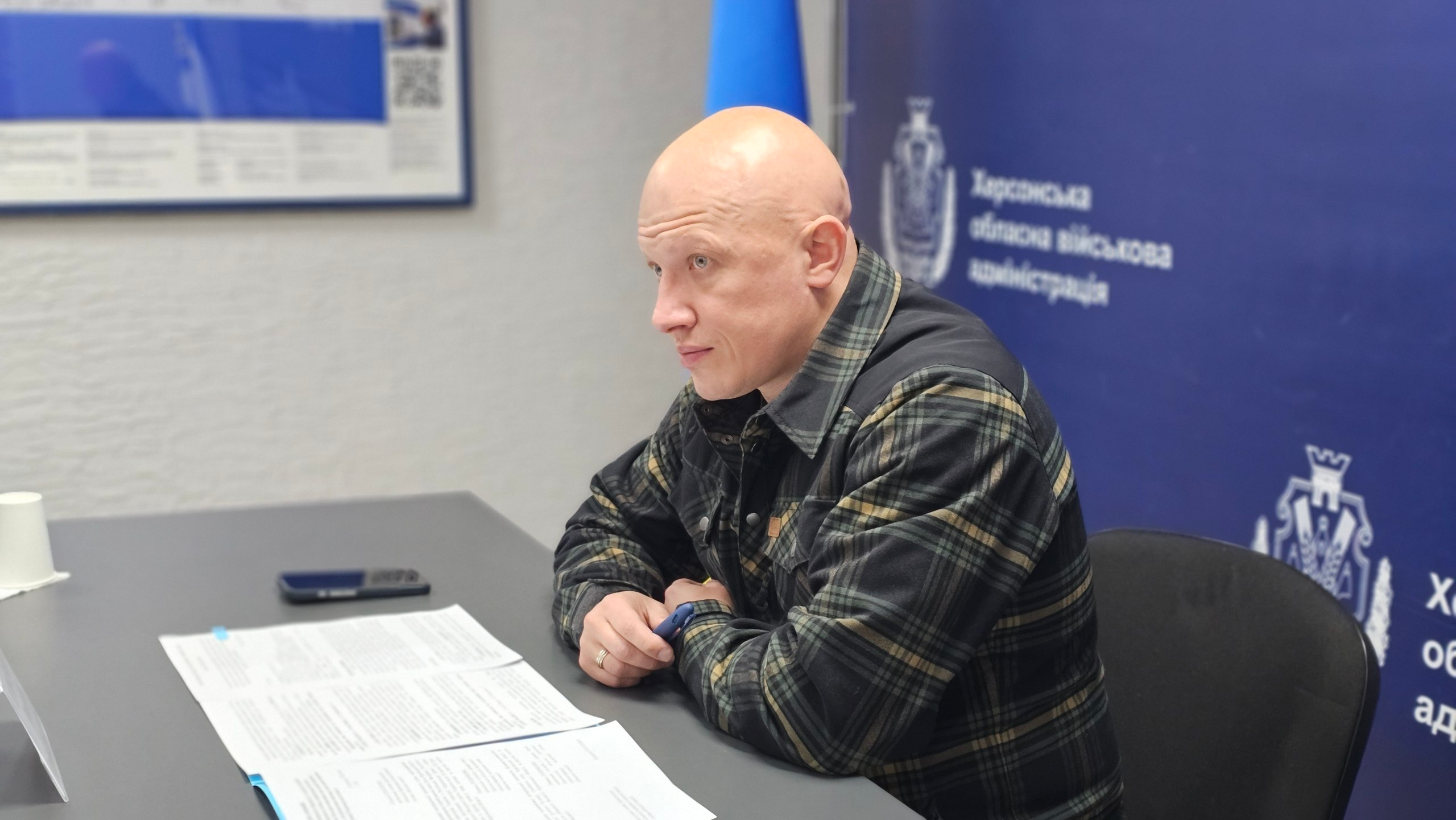 Чоловік у картатій сорочці сидить за робочим столом із документами на фоні синього банера з офіційною символікою.
