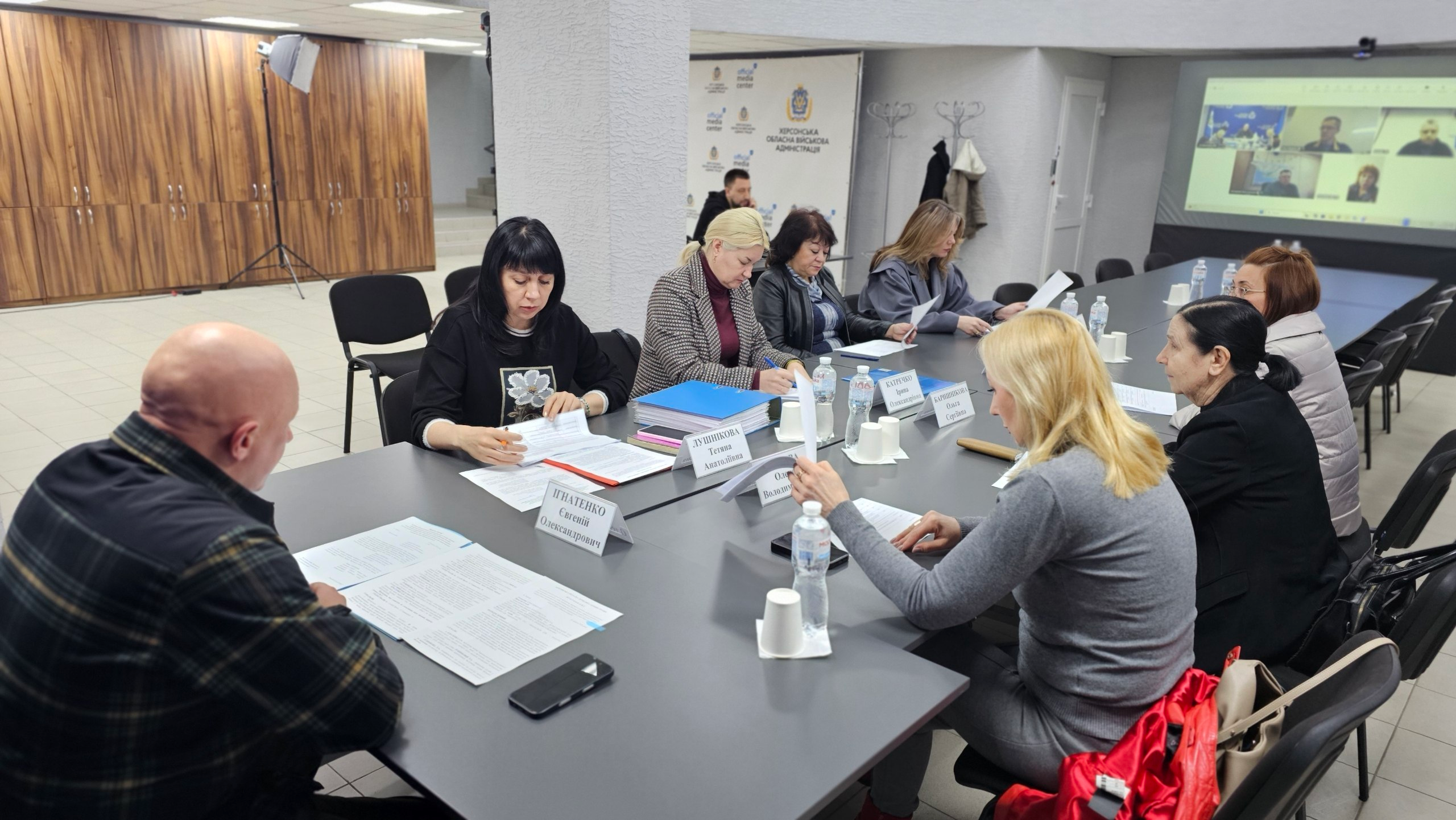Нарада в офісному приміщенні: учасники сидять за довгим столом, працюючи з документами. На задньому плані видно екран із відеозв’язком та банер з офіційною символікою.