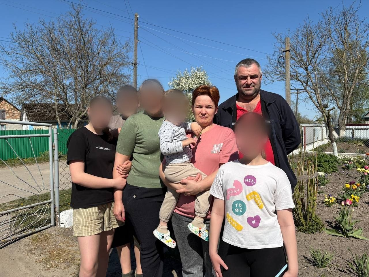 На груповому фото зображена родина на подвір'ї будинку. У центрі стоїть жінка з маленькою дитиною на руках, а поруч із нею — чоловік. Також на знімку присутні четверо дітей, обличчя яких приховано.