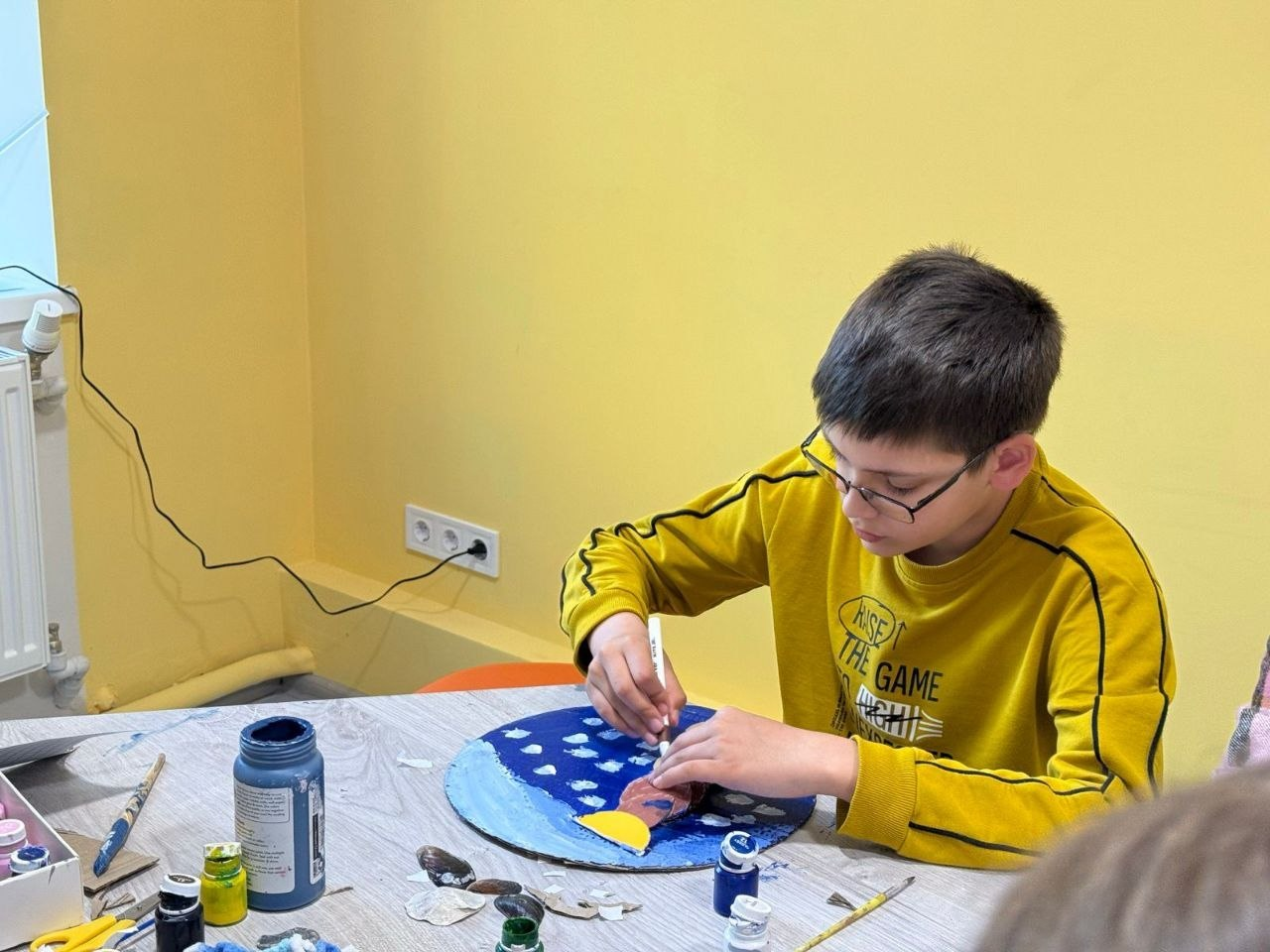 Хлопець у жовтому світшоті зосереджено працює над виробом за столом. Він розфарбовує круглу картонну основу синього кольору, використовуючи фарби та пензлики. На столі навколо нього розкладені баночки з гуашшю та художнє приладдя.