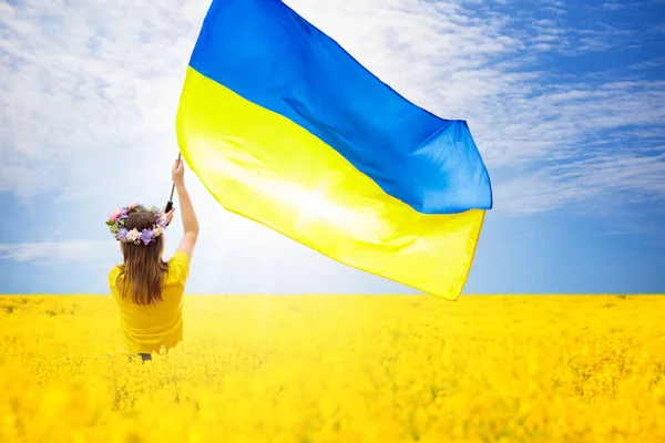 Дівчина у вінку та жовтій футболці стоїть посеред яскраво-жовтого поля, тримаючи великий державний прапор України, що майорить на вітрі. На задньому плані — чисте блакитне небо з легкими хмаринками, а все зображення пронизане сонячним світлом.