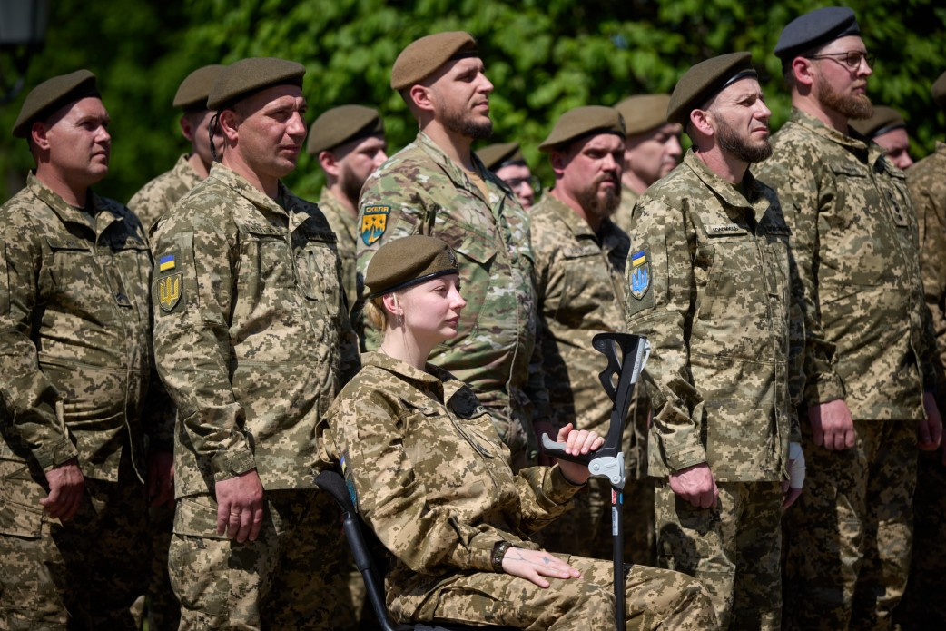 Група військовослужбовців у камуфляжній формі та беретах стоїть у строю просто неба. На передньому плані сидить жінка-військова, яка тримає милицю. На рукавах одностроїв видно нашивки з українською символікою.