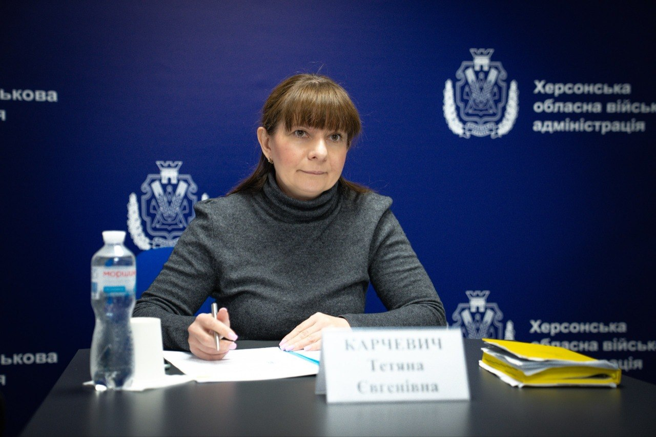 Жінка в сірому светрі з високим коміром сидить за столом на фоні темно-синього банера з офіційною символікою. Вона тримає ручку та дивиться в камеру, перед нею лежать папери та табличка з ім'ям.
