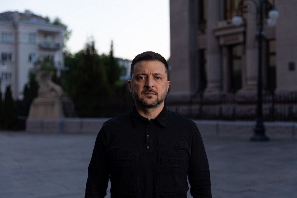Чоловік у чорній сорочці стоїть на вулиці на фоні адміністративної будівлі у сутінках. Він дивиться прямо в камеру з серйозним виразом обличчя.