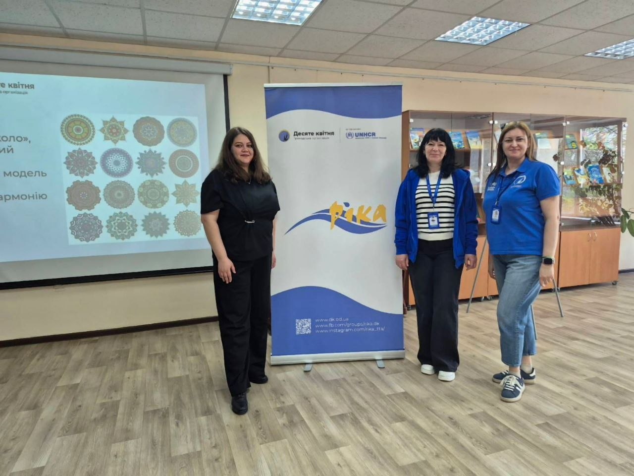 Три жінки стоять у приміщенні біля банера ГО «Десяте квітня» та УВКБ ООН. Поруч розташований проєкційний екран із презентацією візерунків-мандал.