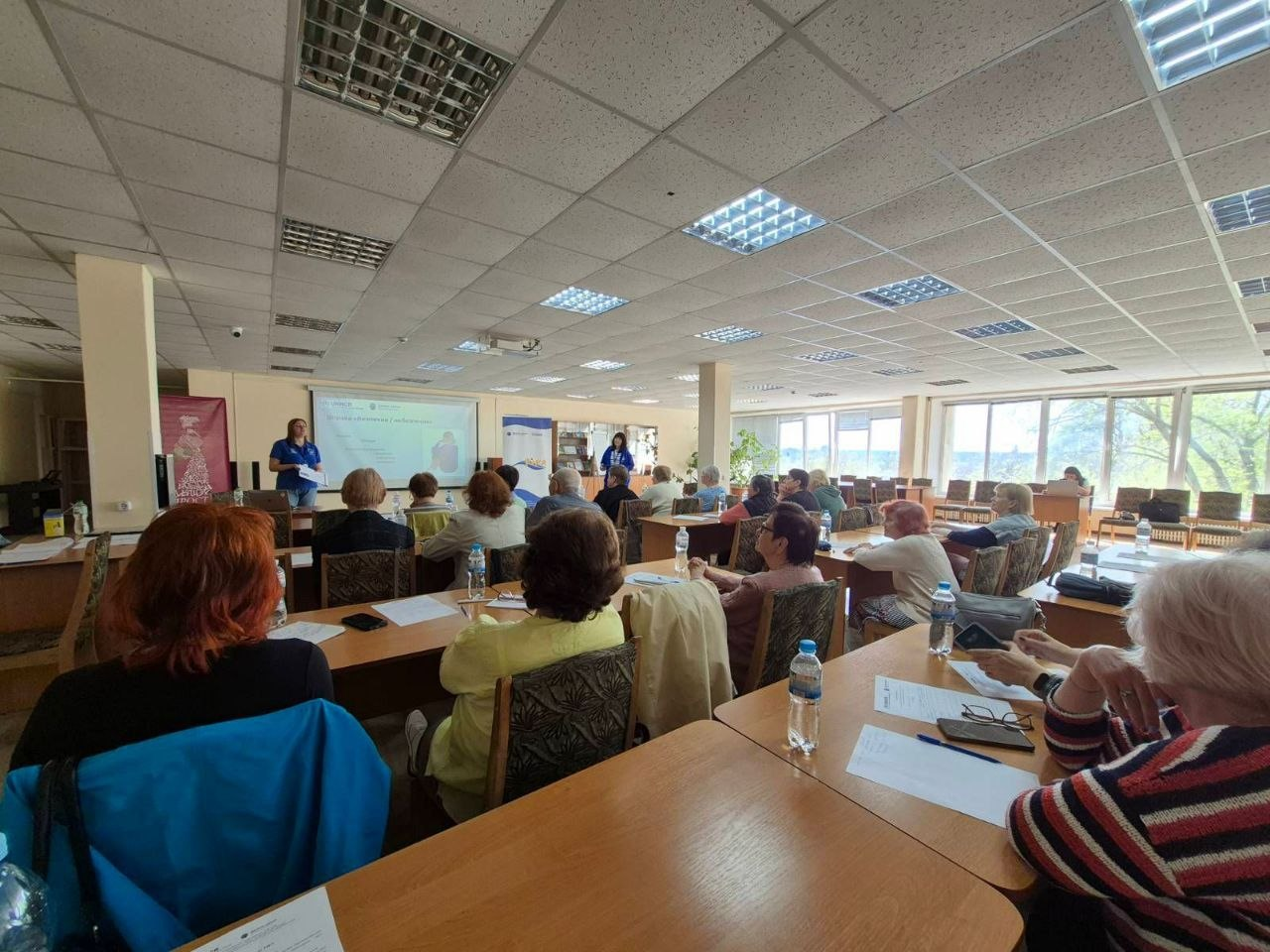 Учасники заходу сидять у залі за столами під час презентації. Спікерки стоять попереду біля екрана та інформаційного банера ГО «Десяте квітня» та УВКБ ООН.