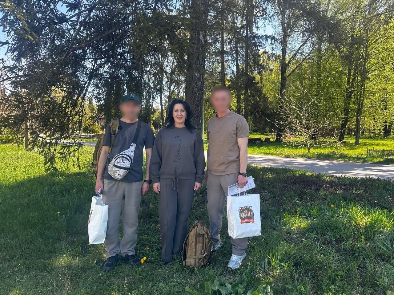 Жінка та двоє чоловіків стоять у парку на фоні дерев. Чоловіки тримають білі паперові пакети, а поруч на траві стоїть військовий рюкзак.