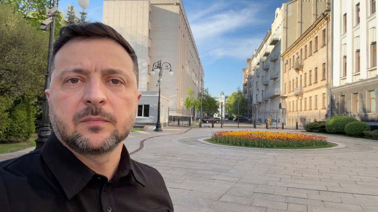 Селфі-знімок чоловіка у чорній сорочці на фоні міської площі з бруківкою та квітковою клумбою. На задньому плані видно багатоповерхові будинки, дерева та освітлювальні ліхтарі під ясним небом.