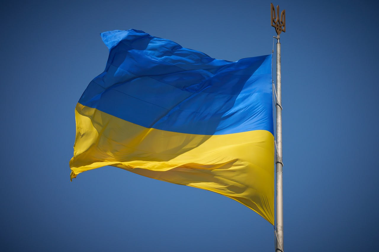 Державний прапор України, що майорить на високому флагштоку на фоні чистого блакитного неба. Верхівка флагштока прикрашена тризубом.