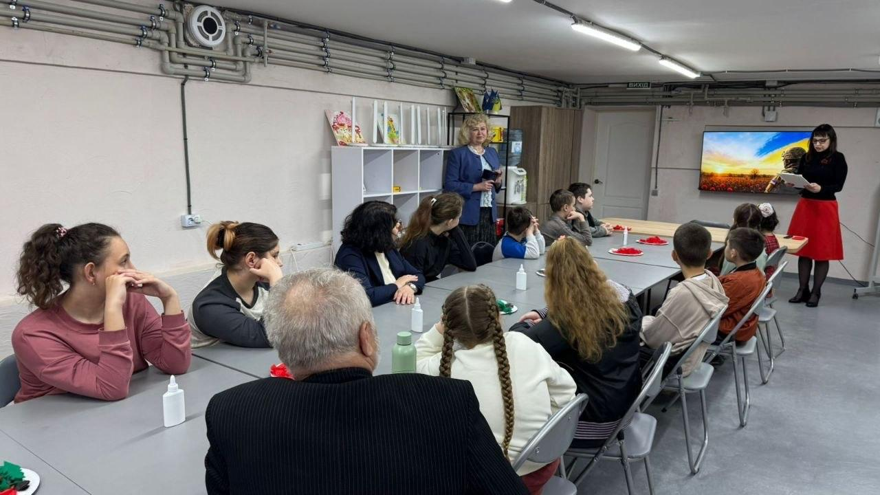 Учасники заходу сидять за столами у приміщенні під час презентації. Попереду стоять спікерки, одна з яких тримає промову, а інша — планшет на фоні екрана з тематичним зображенням.