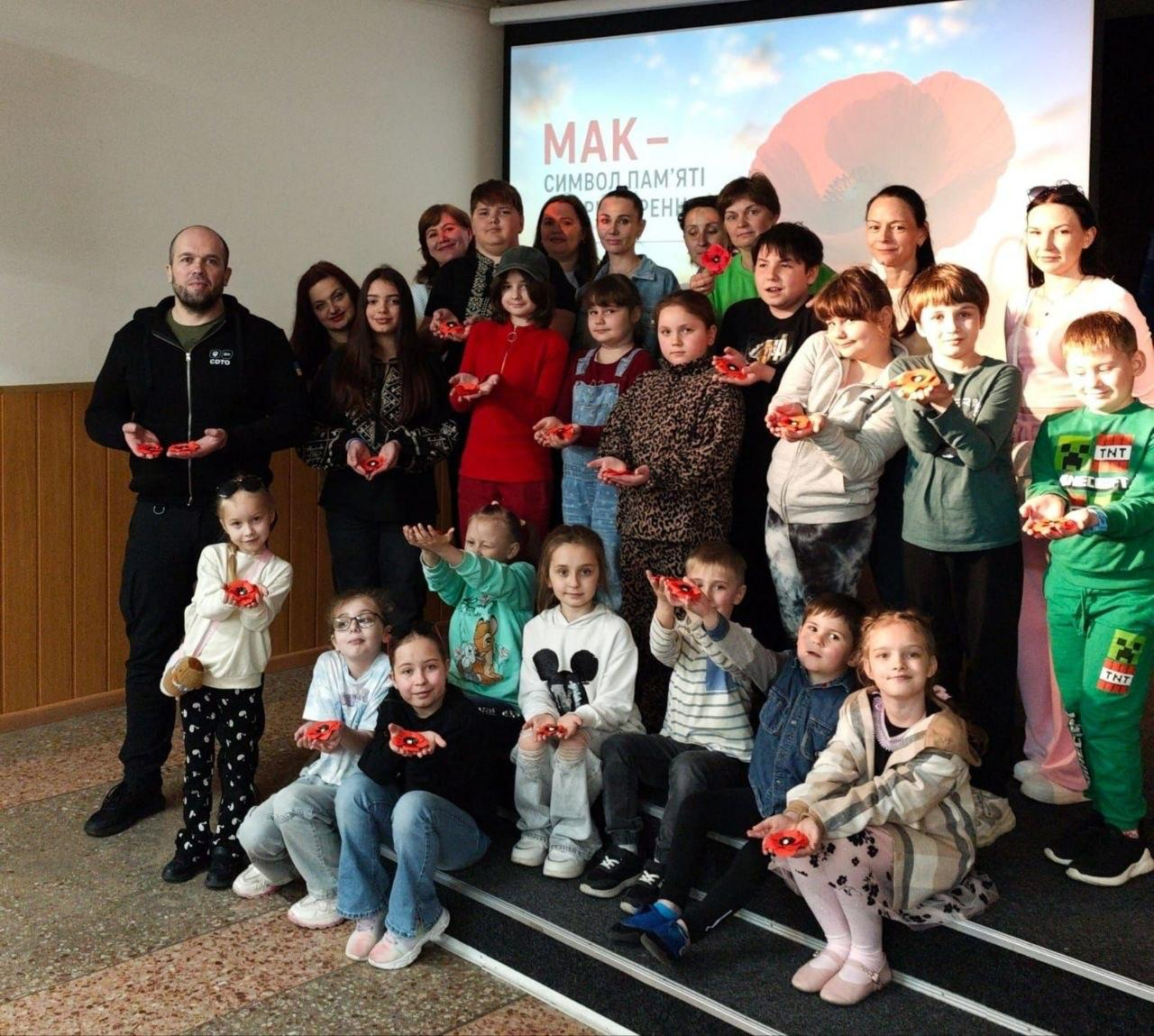 Групова фотографія дітей та дорослих, які тримають у руках червоні маки. На задньому плані — екран із написом «Мак — символ пам'яті» та зображенням квітки.