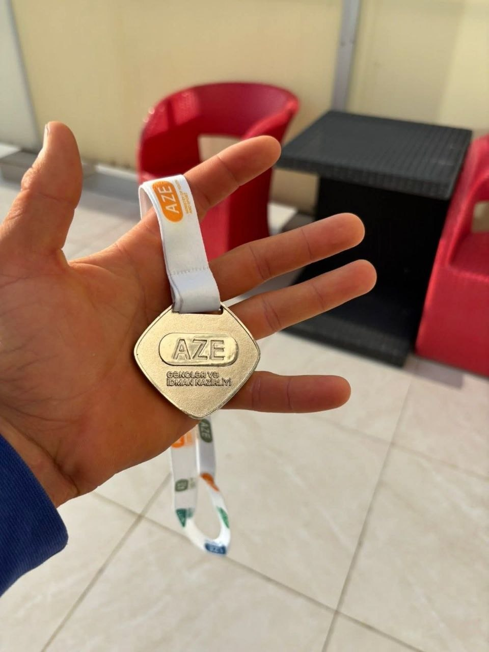 Спортивна медаль ромбоподібної форми з написом «AZE», яку людина тримає в руці. Медаль на білій стрічці з логотипами, ймовірно, отримана за досягнення на міжнародних змаганнях в Азербайджані.
