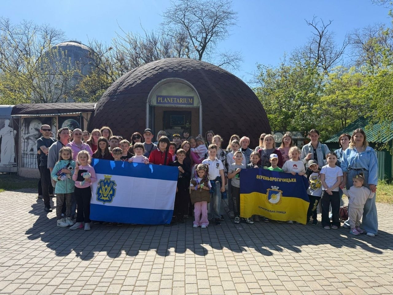 Група дітей та дорослих стоїть на вулиці перед будівлею планетарію. Учасники тримають прапори Херсонської області та Верхньорогачицької територіальної громади, позуючи для спільного фото під час екскурсії.