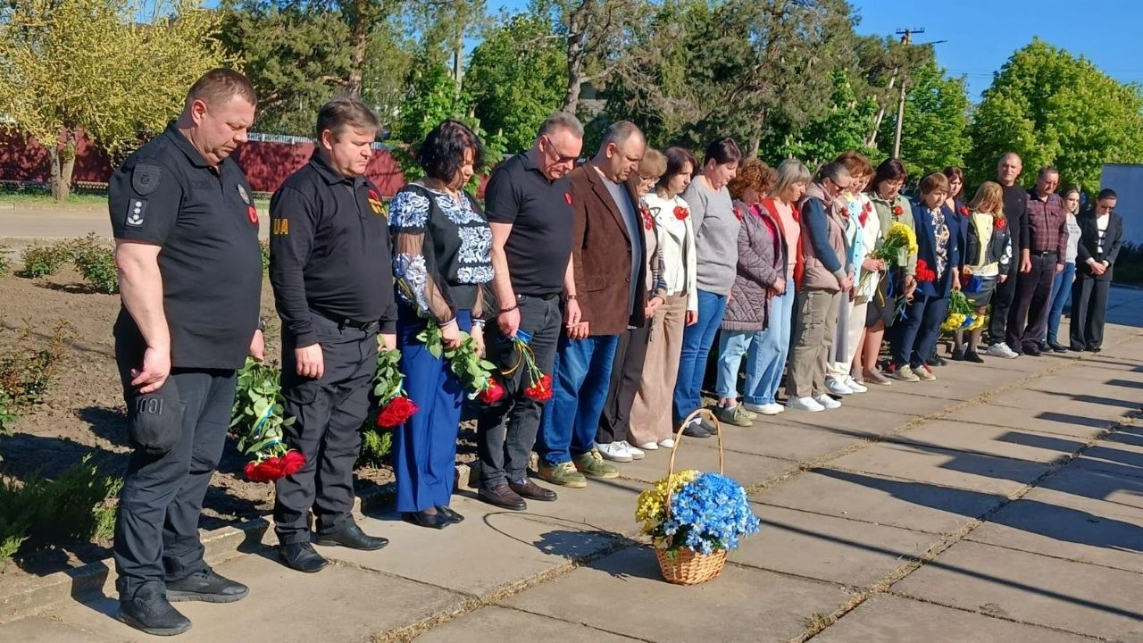 Група людей стоїть у шерензі з похиленими головами під час хвилини мовчання. У руках учасників — червоні троянди, а перед ними на тротуарі стоїть кошик із синьо-жовтими квітами.