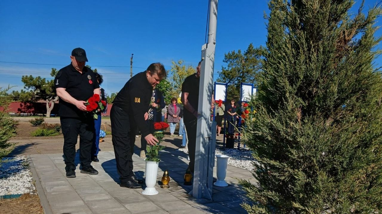 Чоловіки у темному одязі покладають червоні троянди у білу вазу біля підніжжя флагштока. На задньому плані видно інших учасників меморіального заходу та зелені насадження під ясним небом.