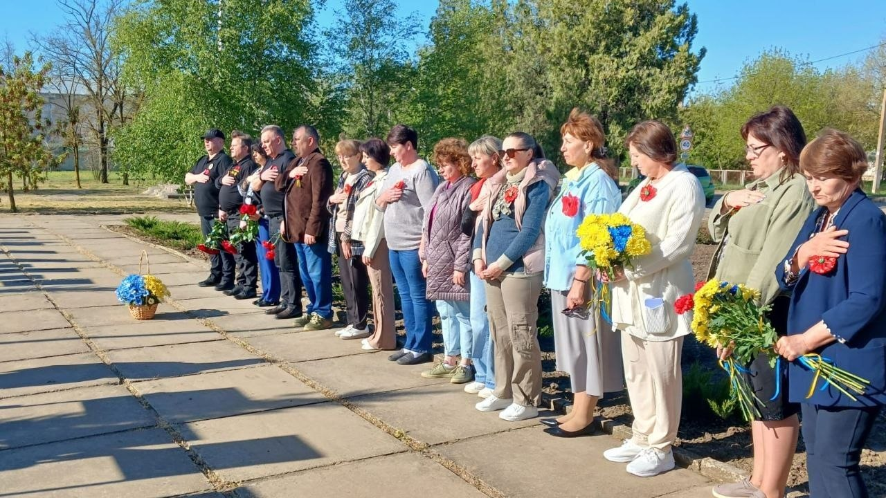 Група людей стоїть у шерензі на вулиці, приклавши праву руку до серця під час урочистого заходу. Учасники тримають квіти, зокрема синьо-жовті букети та червоні маки, а поруч на доріжці стоїть кошик із квітами.