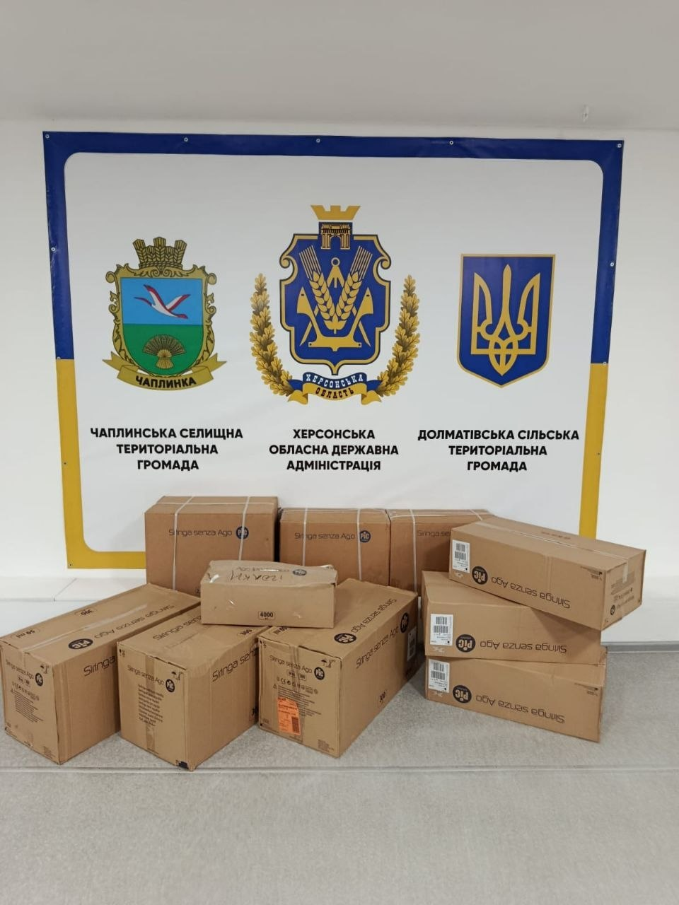 Інформаційний банер із гербами та назвами Херсонської ОДА, Чаплинської та Долматівської громад. На фоні банера на підлозі складені картонні коробки з гуманітарною допомогою.