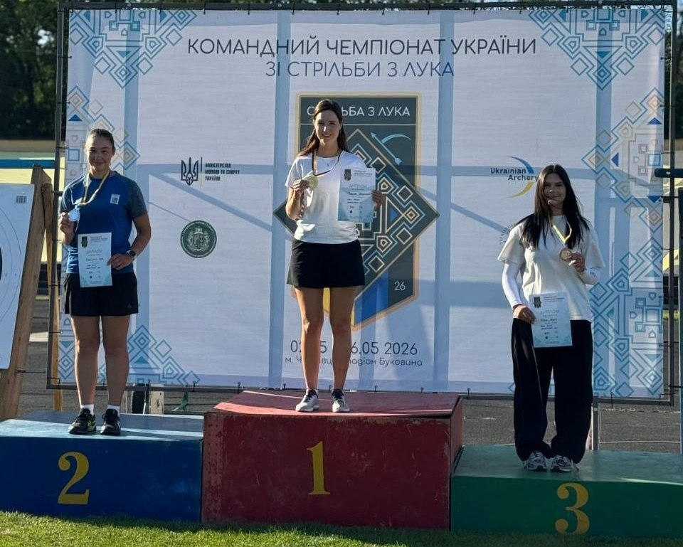 Три спортсменки стоять на п'єдесталі пошани з медалями та дипломами під час церемонії нагородження Командного чемпіонату України зі стрільби з лука. Позаду них — офіційний банер змагань із державною символікою та логотипами організаторів.