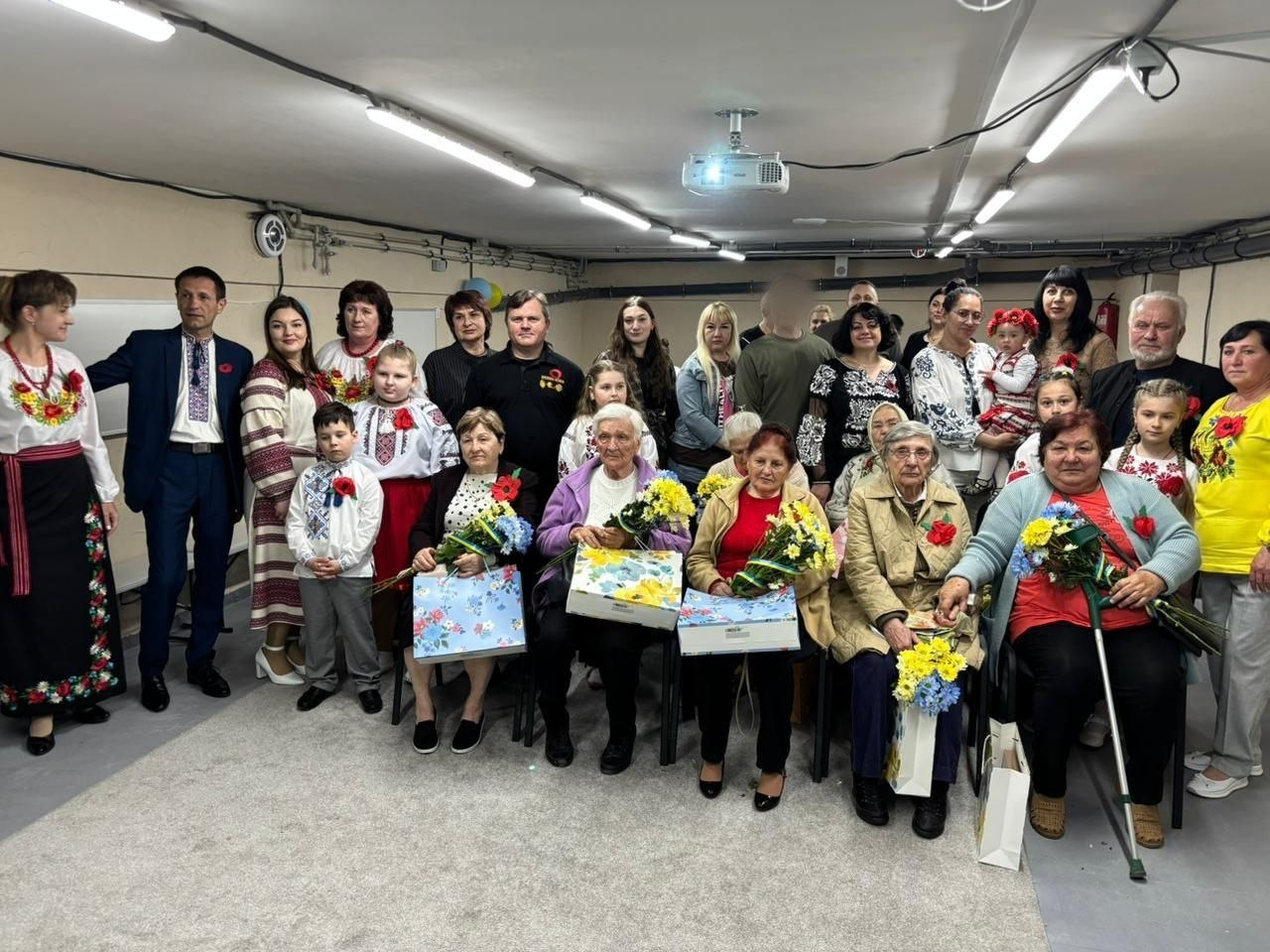 Групова фотографія людей різного віку в приміщенні. Дехто одягнений у вишиванки, жінки старшого віку сидять попереду з букетами квітів та подарунками, прикрашеними синьо-жовтими стрічками.