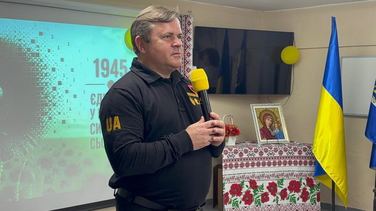 Чоловік у чорному тактичному одязі виступає з мікрофоном у приміщенні. Поруч — державний прапор України, ікона на столі з вишитим рушником та екран із тематичною презентацією до Дня пам'яті.