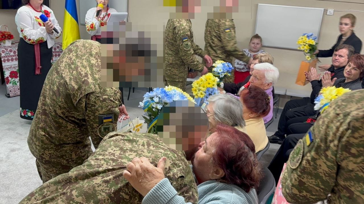 Військовослужбовці вітають жінок старшого віку під час урочистого заходу. Військові вручають синьо-жовті букети та подарунки, а на фоні жінки у вишиванках ведуть програму біля державного прапора.