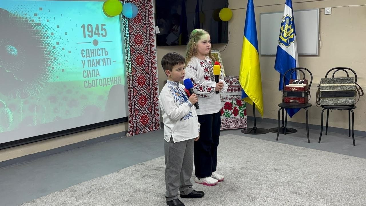 Двоє дітей у вишиванках виступають із мікрофонами під час заходу. Поруч — екран із написом «Єдність у пам'яті — сила сьогодення», державний прапор та музичні інструменти.