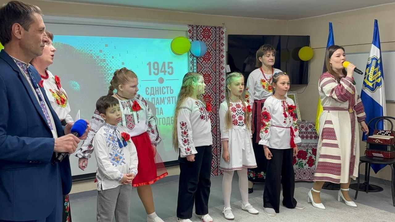Група дорослих та дітей у вишиванках співають під час урочистого заходу. На фоні — проєкційний екран із тематичним написом «Єдність у пам'яті — сила сьогодення», державна символіка та вишитий рушник.