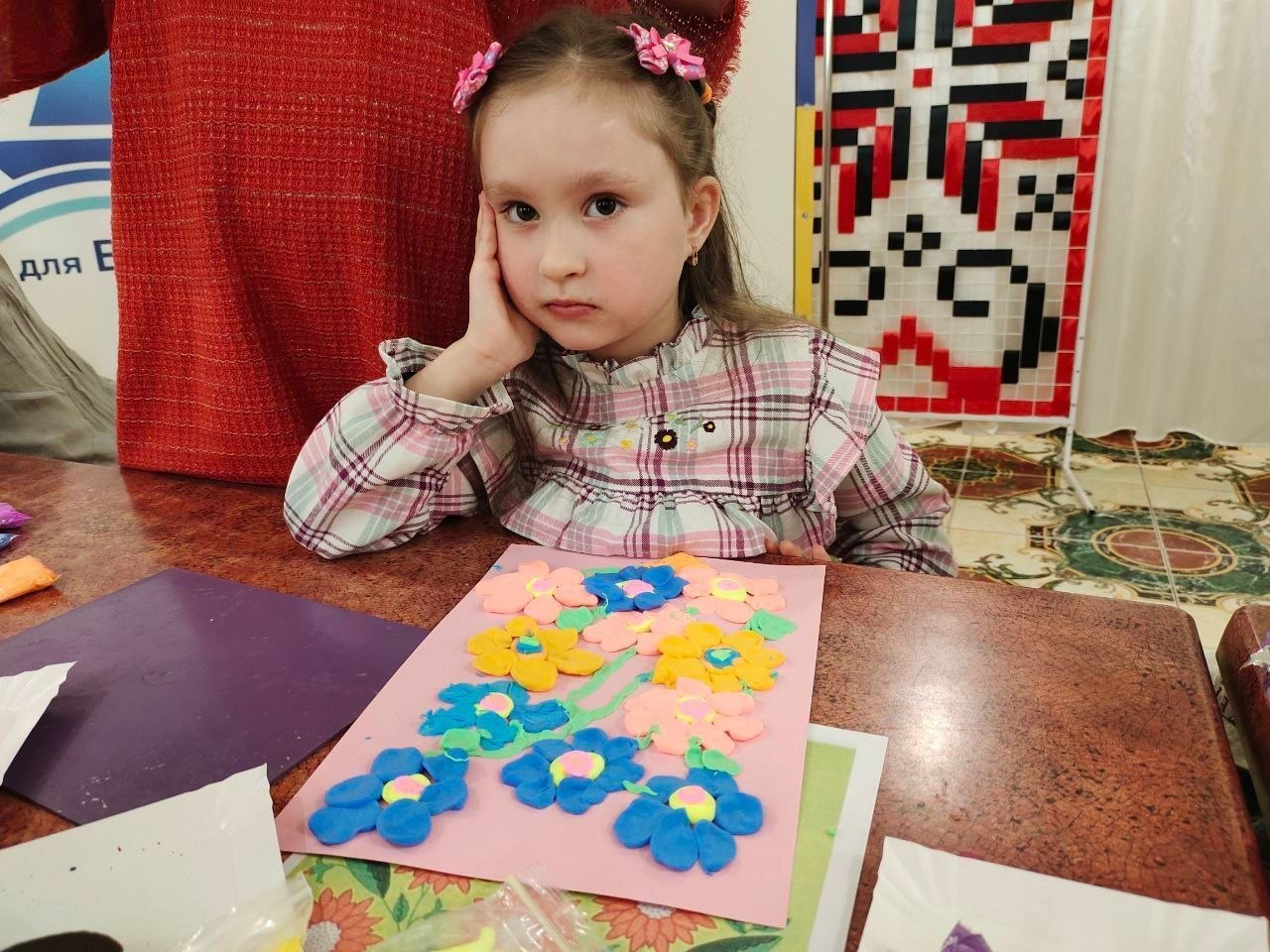 Дівчинка сидить за столом і демонструє свою творчу роботу — аплікацію з яскравих пластилінових квітів на рожевому папері. На задньому плані видно декоративне панно з традиційним українським орнаментом.