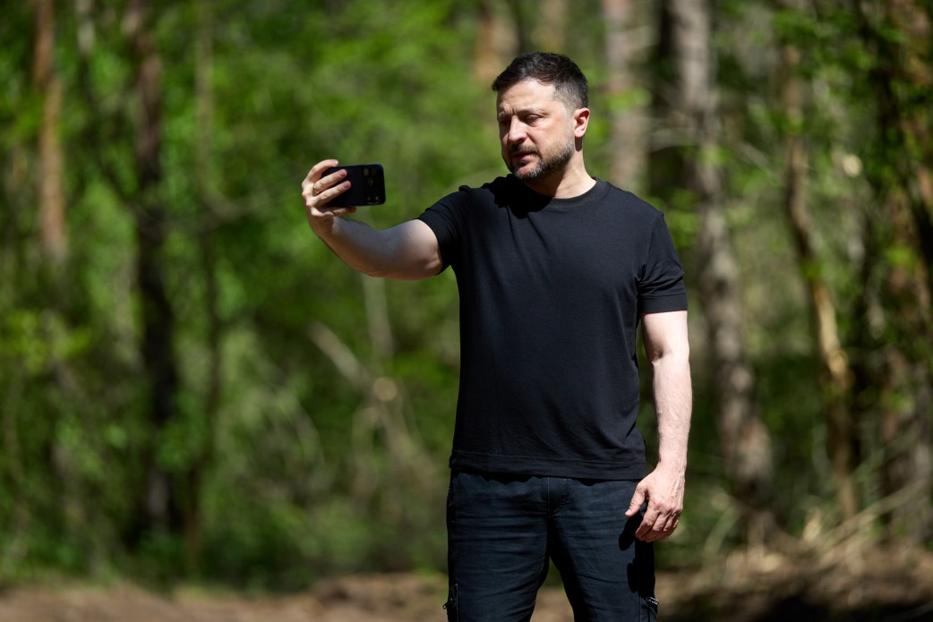 Чоловік у чорній футболці та штанах робить селфі або записує відео на смартфон, тримаючи його на витягнутій руці. Він стоїть на вулиці на фоні розмитих зелених дерев у сонячний день.