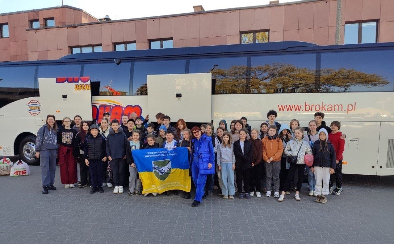 Група дітей та дорослих стоїть на вулиці перед великим білим екскурсійним автобусом. Учасники позують для спільного фото, тримаючи попереду синьо-жовтий прапор із символікою.