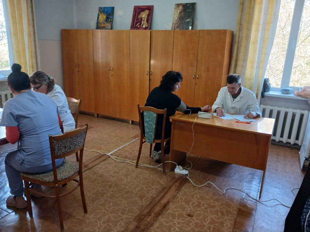 На фото зображено робочий процес у медичному кабінеті. Праворуч за столом медичний працівник у білому халаті вимірює артеріальний тиск пацієнтці та заповнює документи. Ліворуч спиною до камери сидять ще двоє працівників у медичній формі.