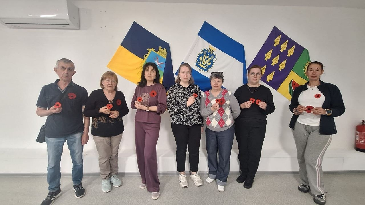 Група людей стоїть у ряд у приміщенні на фоні трьох прапорів. Кожен тримає в руках або має на одязі символічну червону квітку маку. Над людьми на стіні встановлено кондиціонер.