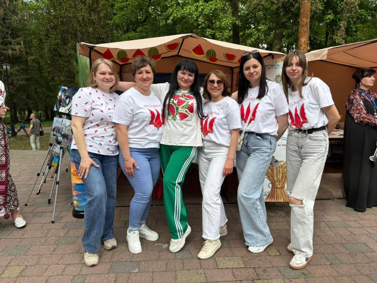 Група з шести жінок усміхається та позує для фото на вулиці перед тентом. Вони одягнені у світлі футболки з патріотичними українськими принтами та малюнками кавунів. На тлі видно дерева та частину ярмаркової локації.