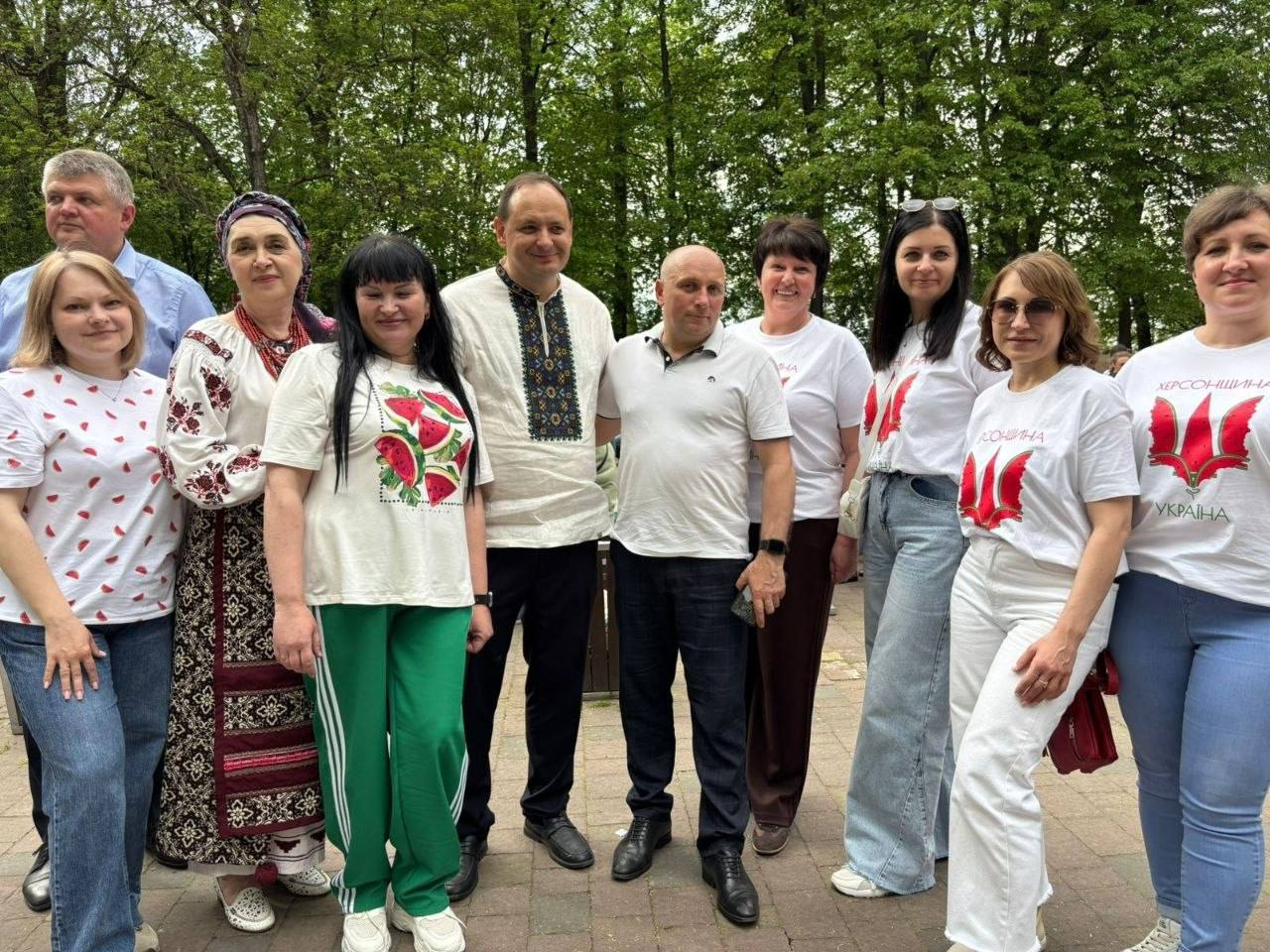 Група людей позує для спільного фото на фоні зелених дерев. У центрі стоять чоловіки у вишиванці та світлій футболці, а поряд з ними — жінки в білих футболках із патріотичними принтами, зображеннями кавунів та традиційному українському вбранні. Усі присутні посміхаються, створюючи доброзичливу атмосферу.