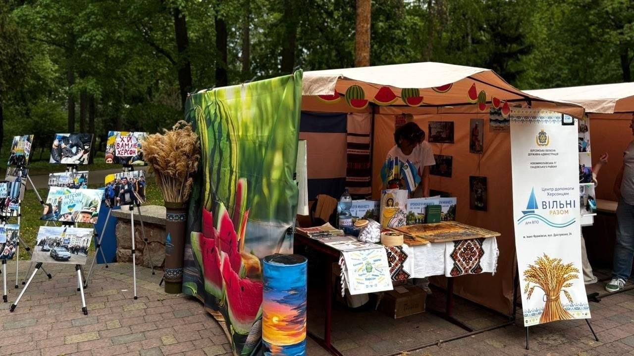 Вулична експозиція в парку з інформаційними стендами та фотовиставкою. Поруч розташований намет, прикрашений декором із кавунами, де на столі з вишитою скатертиною представлені друковані матеріали та символіка. На передньому плані видно банер благодійного центру допомоги херсонцям.