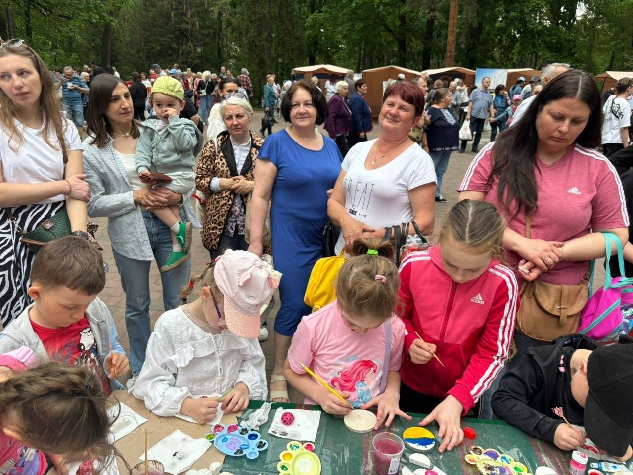 Група дітей та дорослих на масовому заході в парку. На передньому плані діти зосереджено малюють за довгим столом, використовуючи фарби та пензлики. За ними стоять дорослі, які спостерігають за процесом, а на задньому плані видно натовп людей та ярмаркові намети серед дерев.