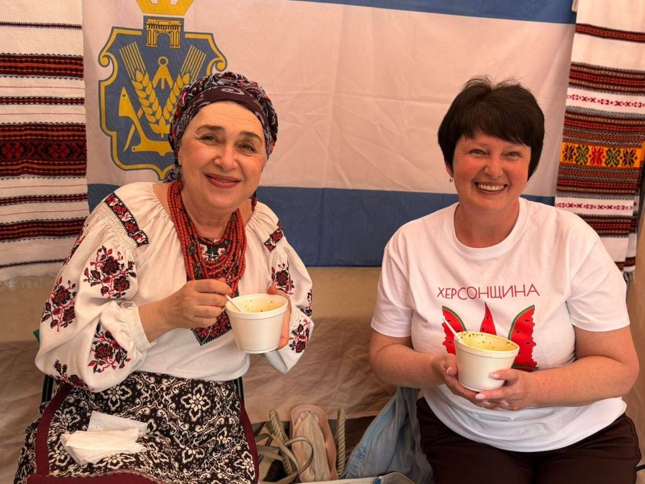Дві усміхнені жінки позують із невеликими білими піалами з їжею. Жінка ліворуч одягнена в традиційну українську вишиванку та квітчасту хустку, а жінка праворуч — у білу футболку з написом «ХЕРСОНЩИНА» та стилізованим зображенням тризуба у вигляді скибок кавуна. На тлі видно прапор із гербом та вишиті рушники.