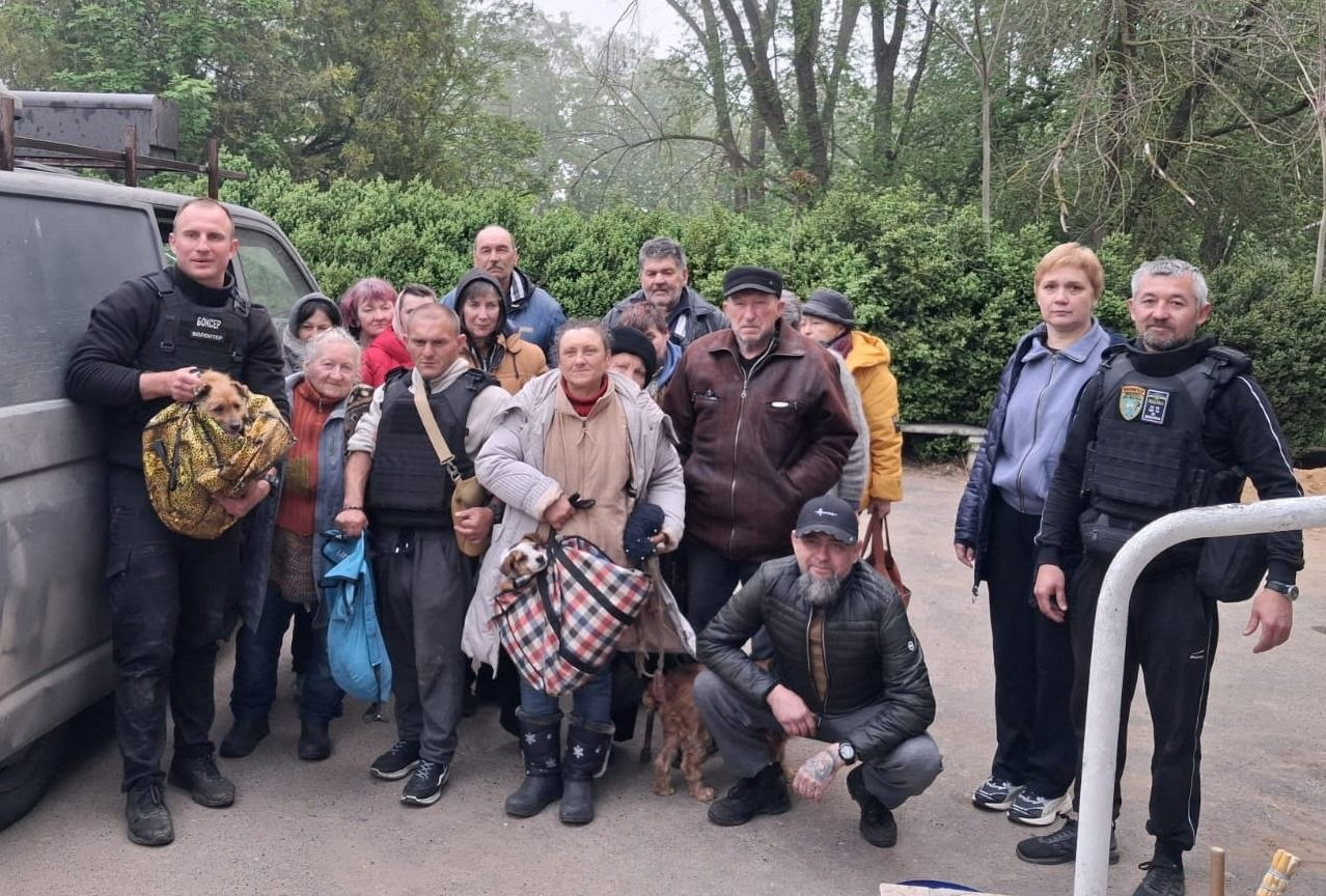 Група людей різного віку стоїть на вулиці поруч із мікроавтобусом. Серед них є чоловіки у захисному спорядженні, один з яких тримає на руках собаку, та люди з сумками-переносками для тварин. На тлі видно густі зелені кущі та дерева, що створює атмосферу зборів або евакуації.