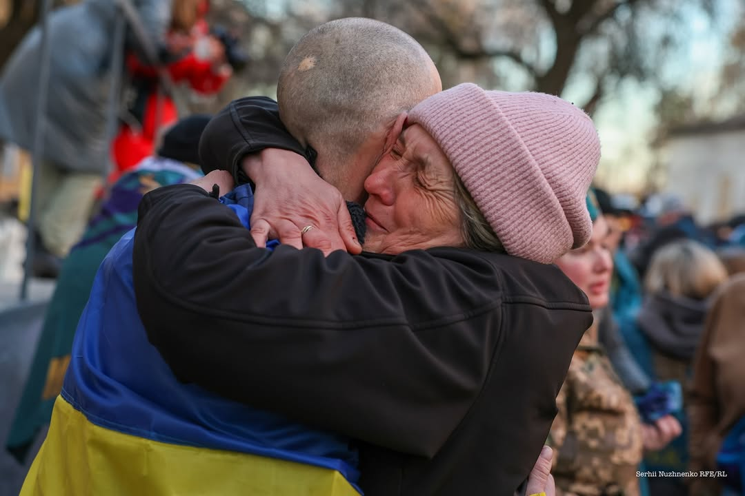 Емоційний момент зустрічі: літня жінка у рожевій в'язаній шапці зі сльозами на очах міцно обіймає за шию чоловіка з голеною головою. На плечах чоловіка закріплений синьо-жовтий прапор України, а на задньому плані розмито видно інших людей і дерева.