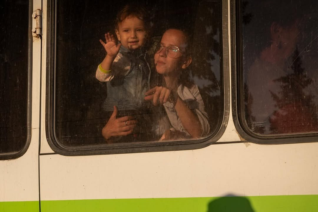 Жінка в окулярах та маленька дитина дивляться на вулицю через вікно білого автобуса з зеленою смугою. Дитина підняла руку, махаючи на прощання, а жінка підтримує її та вказує пальцем уперед.