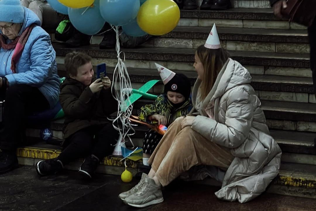 Жінка та двоє дітей у святкових ковпаках сидять на сходах у приміщенні. Поруч із ними видно сині та жовті повітряні кульки, іграшкові літачки, а один із хлопчиків знімає щось на мобільний телефон.