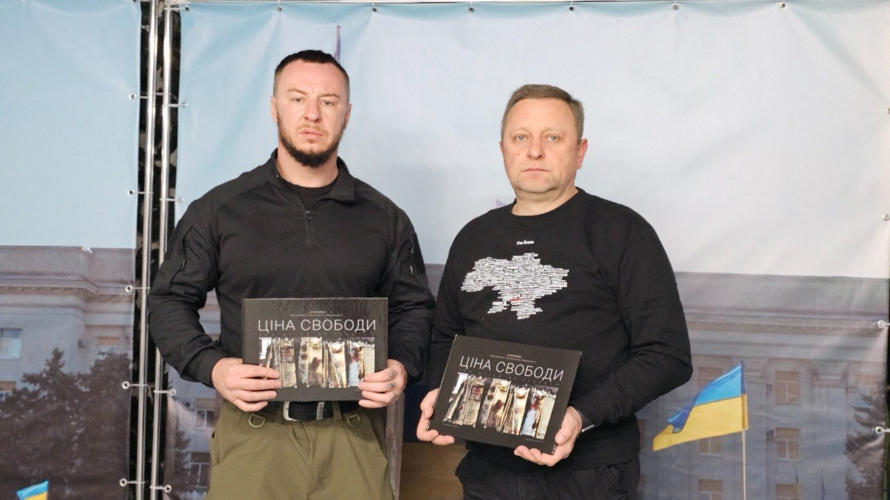 Два чоловіки у темному одязі стоять поруч і тримають у руках примірники книги з написом «Ціна свободи» на обкладинці. На задньому плані видно банер із зображенням будівлі та державного прапора України.