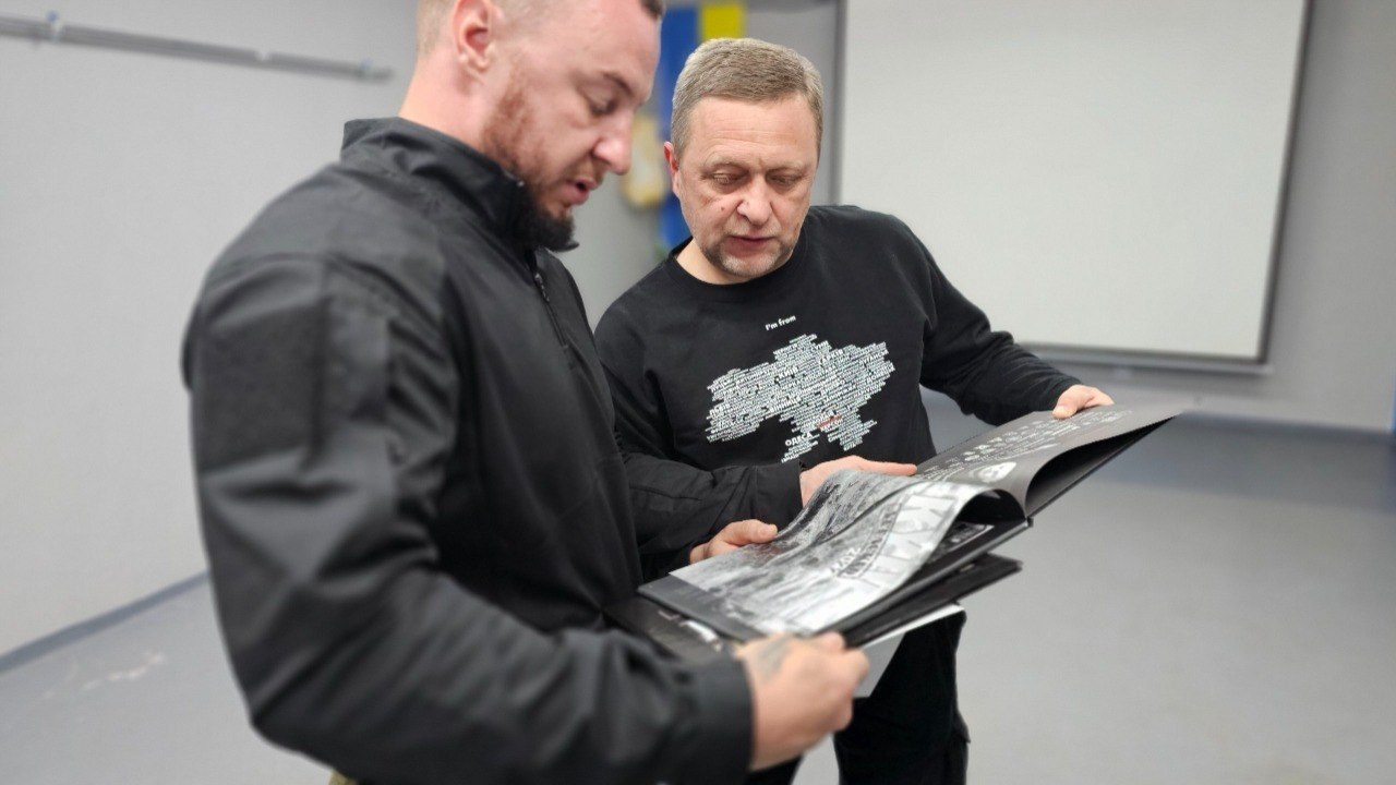 Два чоловіки у чорному одязі стоять поруч у світлій кімнаті та разом розглядають розгорнуту книгу з ілюстраціями. Один із них тримає книгу, а інший схилився над сторінками, уважно вивчаючи вміст.