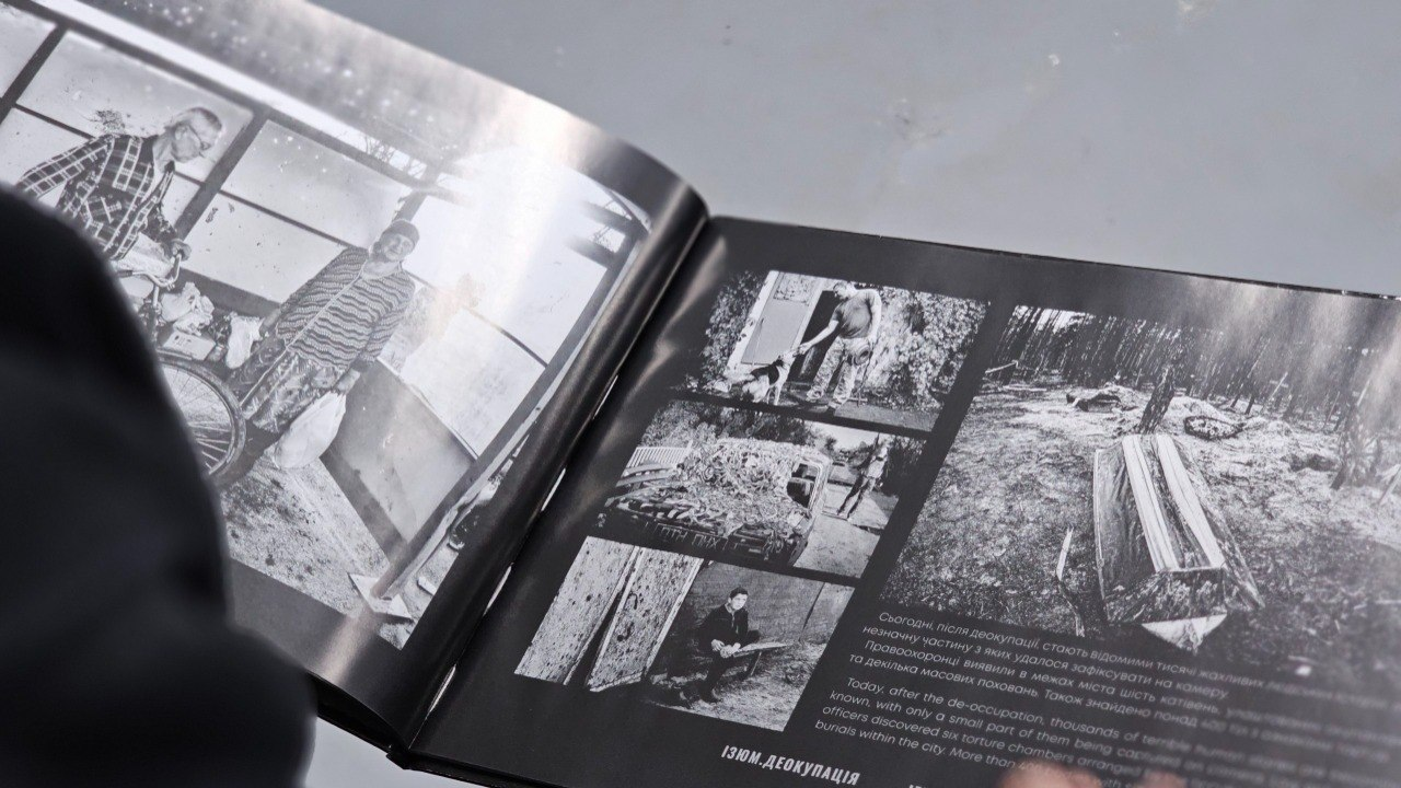 Розгорнута книга у темній обкладинці з чорно-білими фотографіями та текстом двома мовами на сторінках. На знімках зображено лісову місцевість, людей та побутові сцени, а внизу сторінки міститься підпис «ІЗЮМ. ДЕОКУПАЦІЯ».
