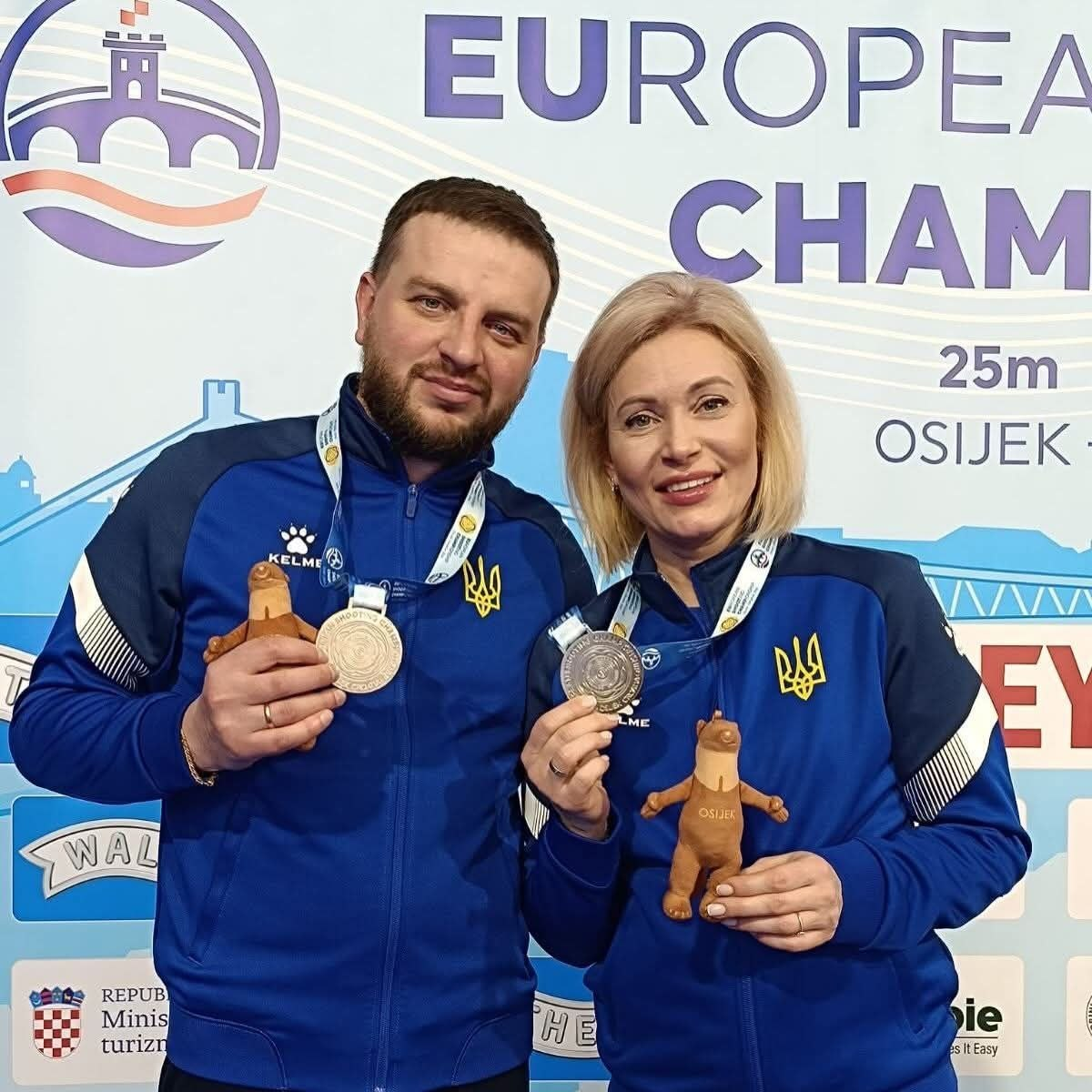 Чоловік та жінка у синій спортивній формі з гербом України стоять поруч і посміхаються. Вони тримають у руках завойовані медалі та невеликі м'які іграшки-талісмани на тлі офіційного банера змагань із написом «EUROPEAN CHAMPIONSHIP 25m OSIJEK».