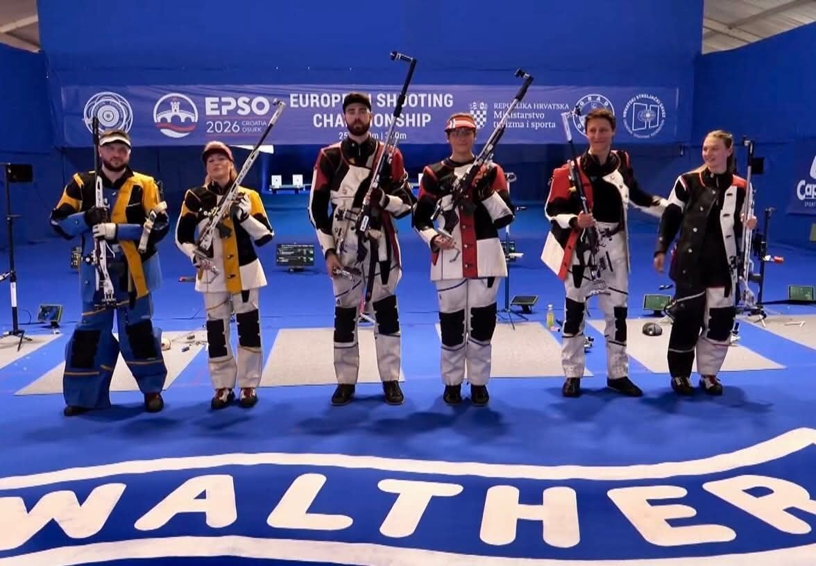 Шість спортсменів у спеціальних стрілецьких костюмах стоять у ряд на синьому покритті стрільбища, тримаючи в руках гвинтівки. На задньому плані розміщено великий банер змагань «EUROPEAN SHOOTING CHAMPIONSHIP», а на передньому плані видно напис «WALTHER».